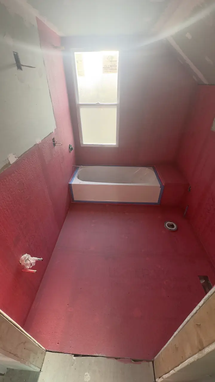 Small bathroom with red waterproofing membrane on the walls and floor, window above the tub.