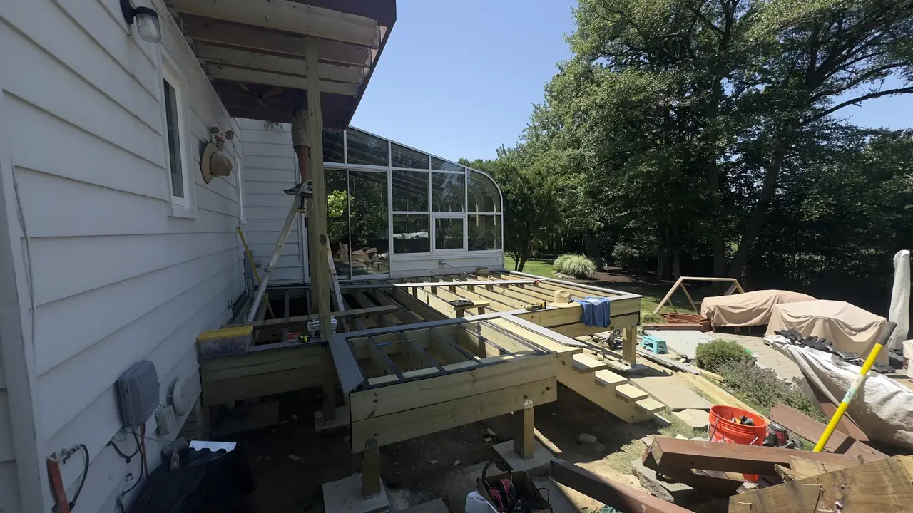 Deck framing meeting a sunroom wall, lumber stacked beside it.